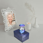 Tiny urn for ashes with blue starry galaxy artwork displayed on a presentation box beside a loved one’s memorial portrait