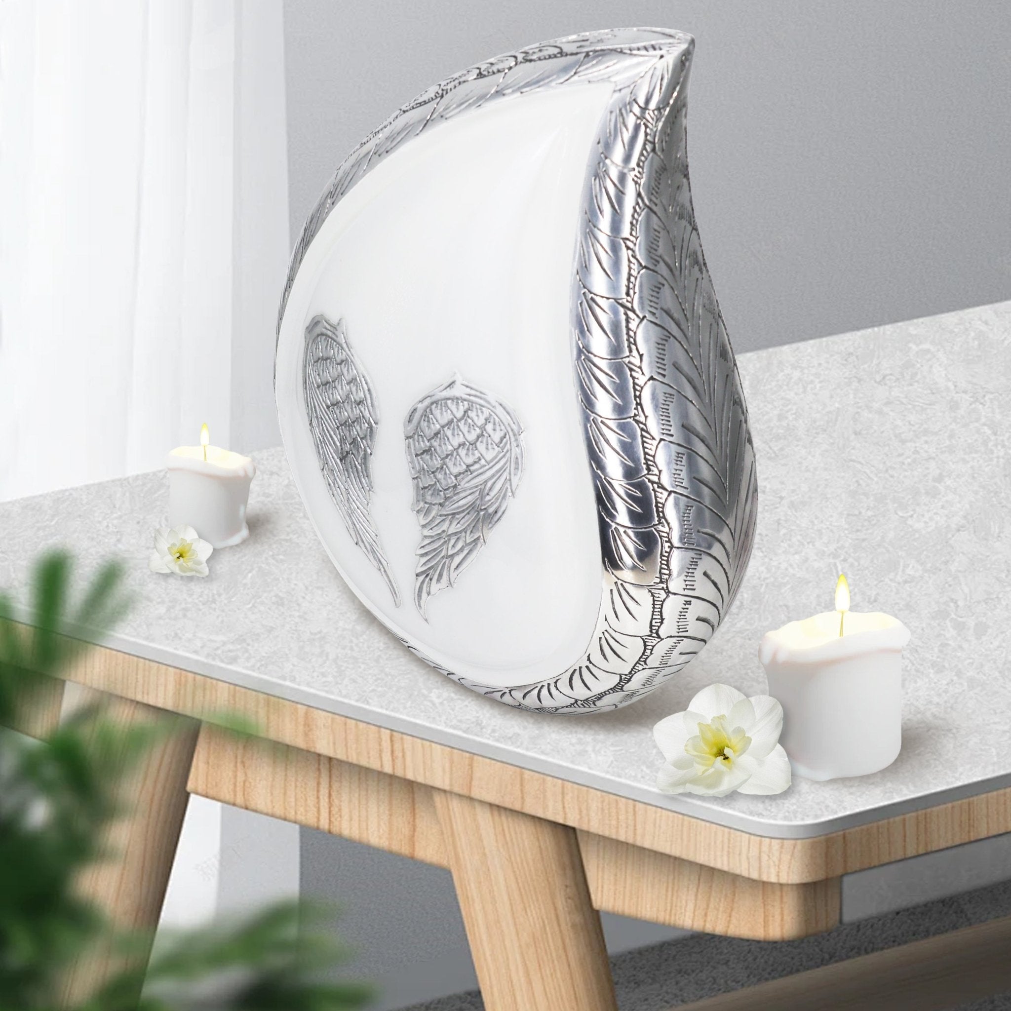 White angel wing teardrop metal burial urns for ashes adults placed on wooden side table with candles for home mantel decor