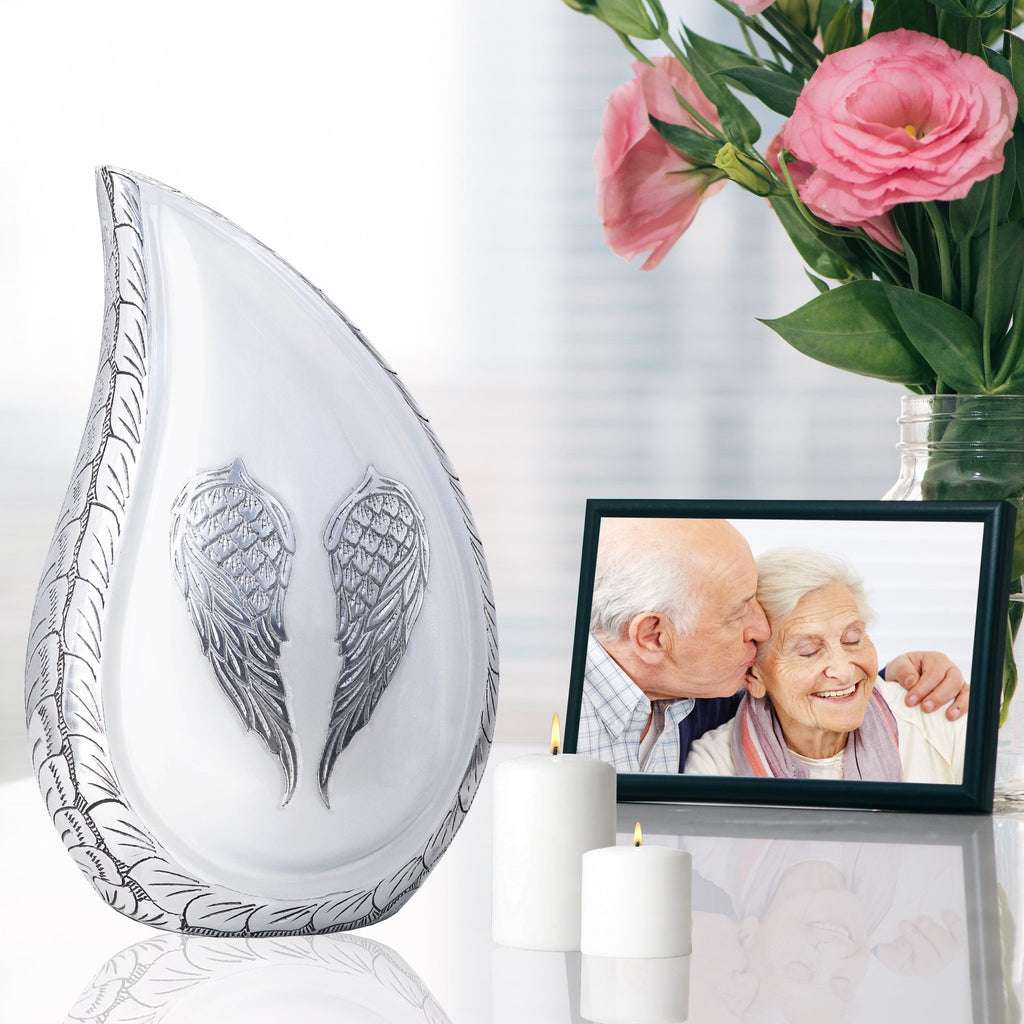 White engraved angel wings teardrop aluminum funeral urn set on reflective table with family photo and pink blooms