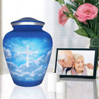 Religious urn for human ashes with blue sky cross artwork displayed beside pink flowers and a framed memorial photo