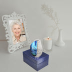 Ocean scenery tiny brass keepsake urn set atop navy gift box beside memorial candle and vintage framed portrait