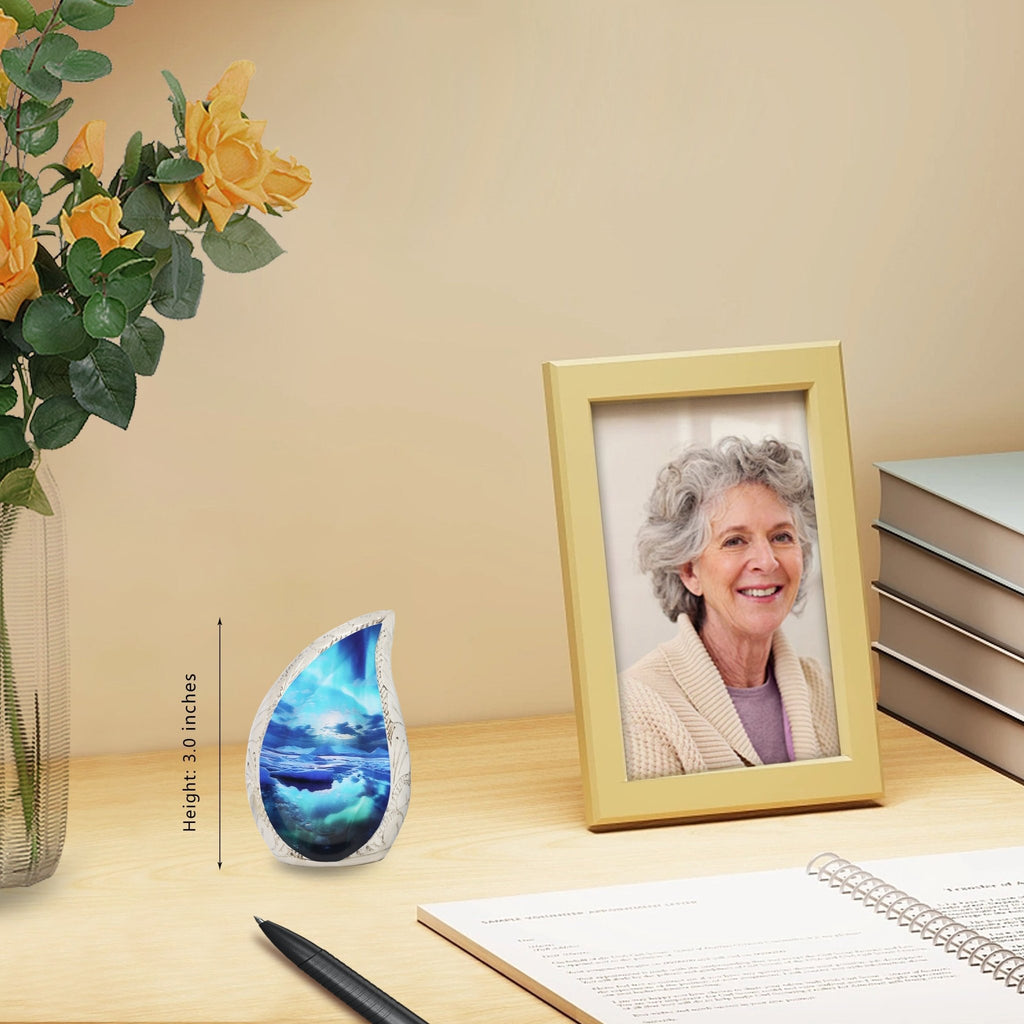 Ocean wave teardrop brass keepsake urn laid on wooden desk paired with yellow roses and framed family portrait