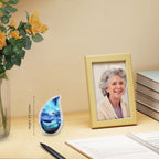 Ocean wave teardrop brass keepsake urn laid on wooden desk paired with yellow roses and framed family portrait