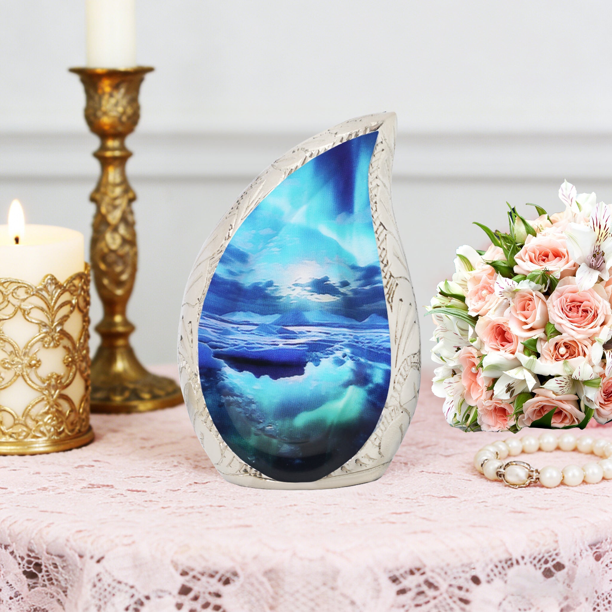Ocean wave brass teardrop ash urn set on lace tablecloth with gold candlesticks and pink funeral rose bouquet