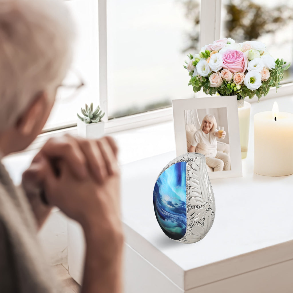 Seascape engraved micro ash urn placed on bright windowsill next to fresh bouquets, candle and loved one photo frame