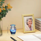 Tiny urn for ashes with turquoise Northern Lights artwork displayed on a desk beside flowers and a family photo
