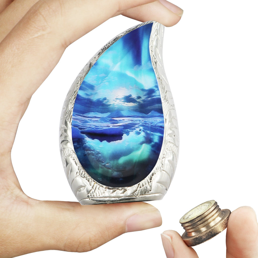 Two hands holding ocean wave printed brass teardrop mini urn and detached threaded bottom lid for human ash storage
