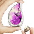 Brass keepsake urn for ashes with purple butterfly teardrop artwork held in hand beside its secure bottom screw lid