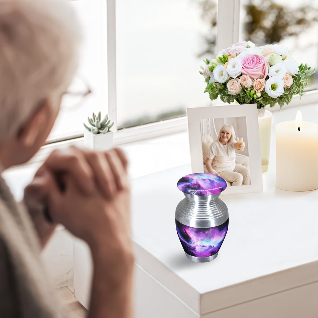 Small urns for human ashes with purple galaxy design displayed near an elderly woman in a quiet home memorial
