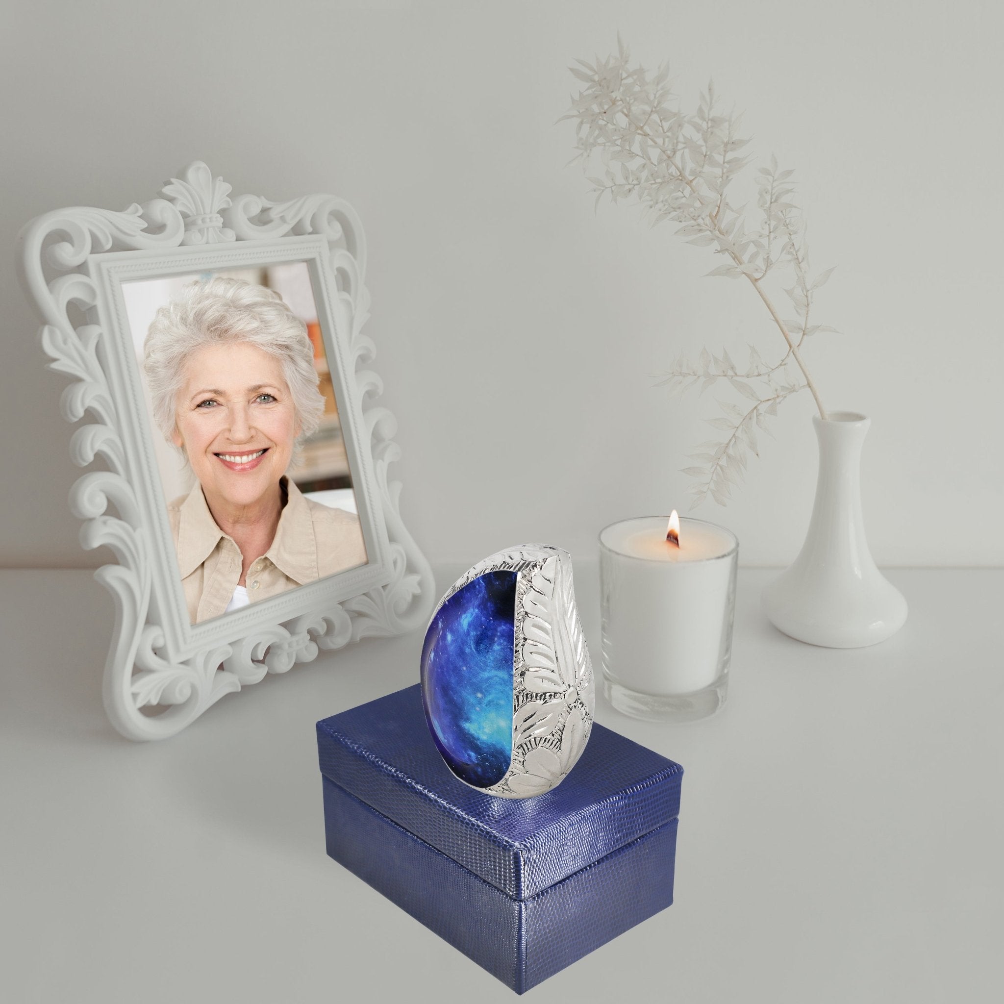 Blue galaxy engraved brass micro cremation urn placed atop gift box with memorial candle and vintage framed portrait