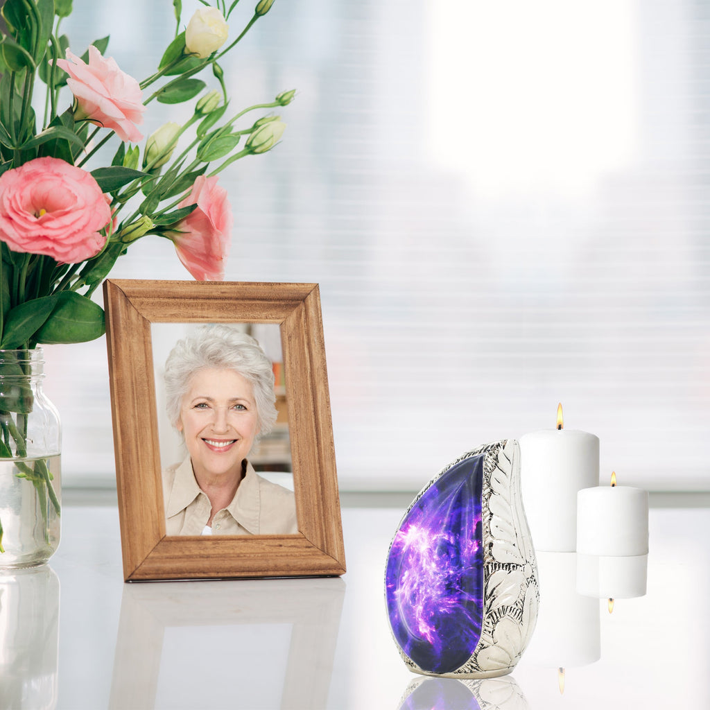 Vintage styled purple nebula brass micro cremation urn displayed on reflective table with pink blooms and memorial portrait