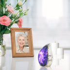 Vintage styled purple nebula brass micro cremation urn displayed on reflective table with pink blooms and memorial portrait